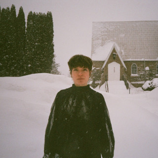 Eine weiße Frau mit kurzen, braunen Haaren und schwarzer Kleidung steht im hohen Schnee vor einer verschneiten Kirche.